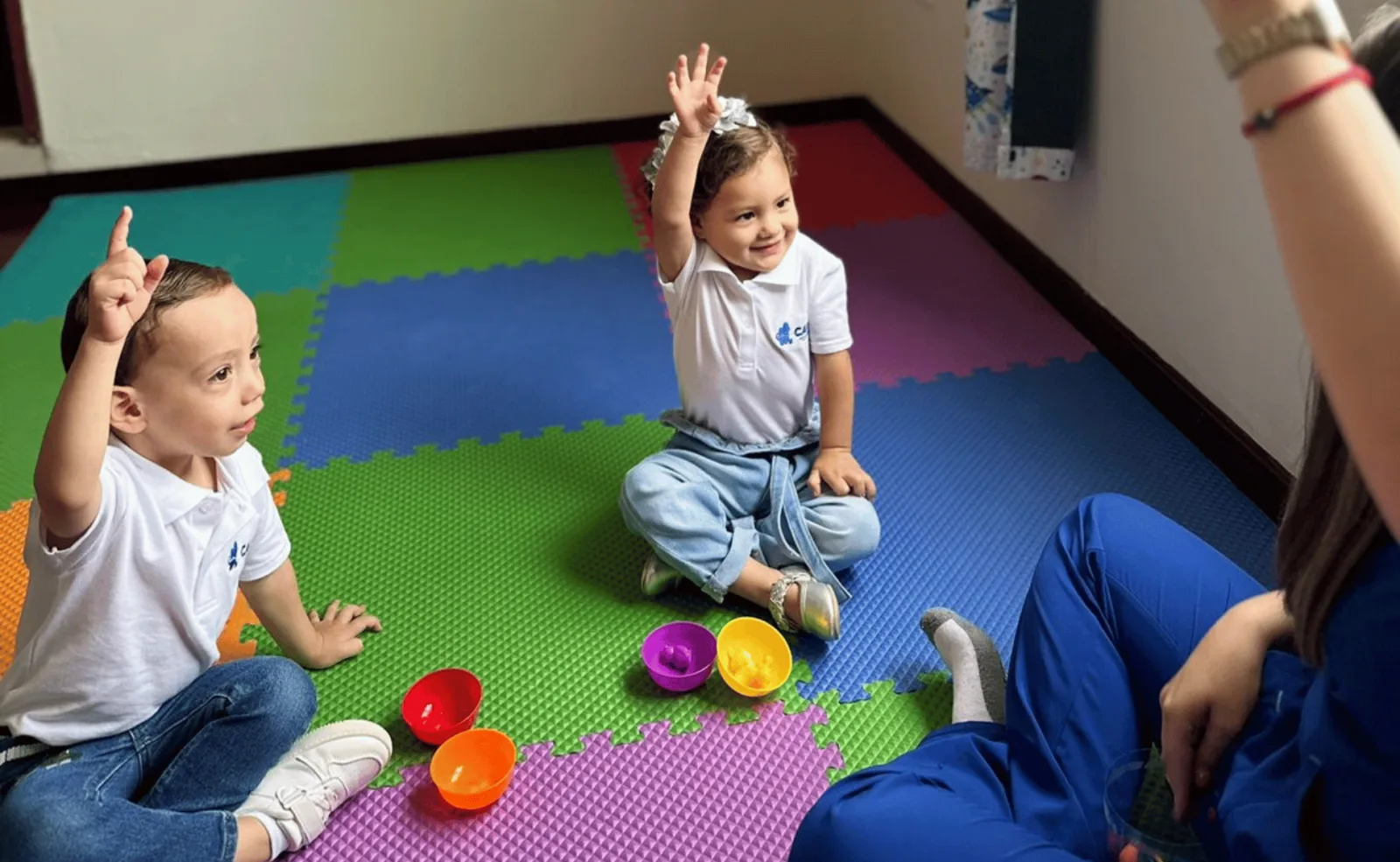 Dos niñas y una adulta levantan la mano durante una actividad sobre tapetes de colores