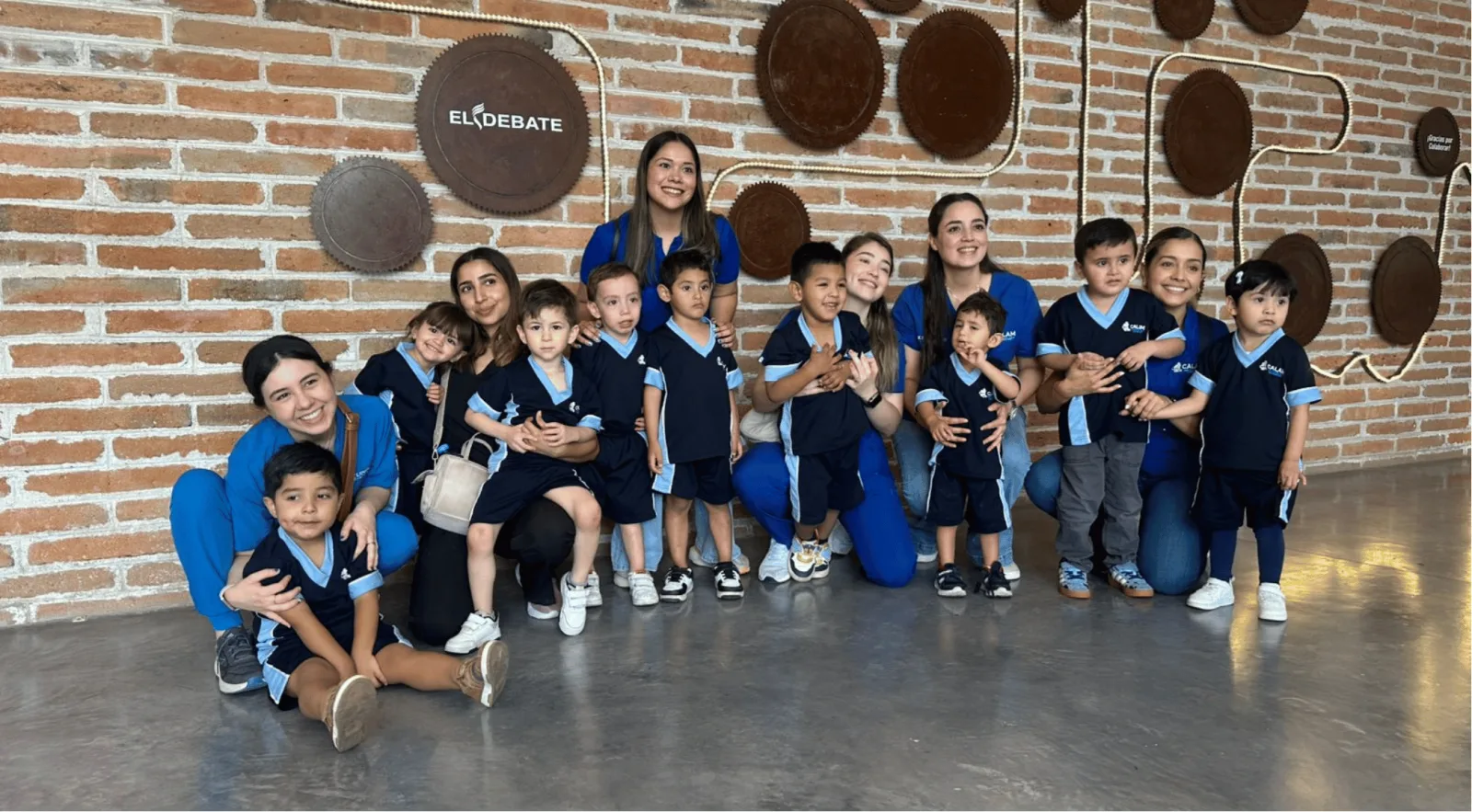 Niñas, niños y adultas con uniformes de CALAM frente a un muro de ladrillo