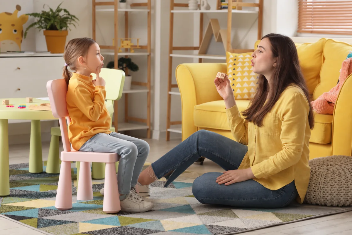 Profesional y niña sentadas frente a frente, modelando un sonido mientras la adulta sostiene un bloque de letras.