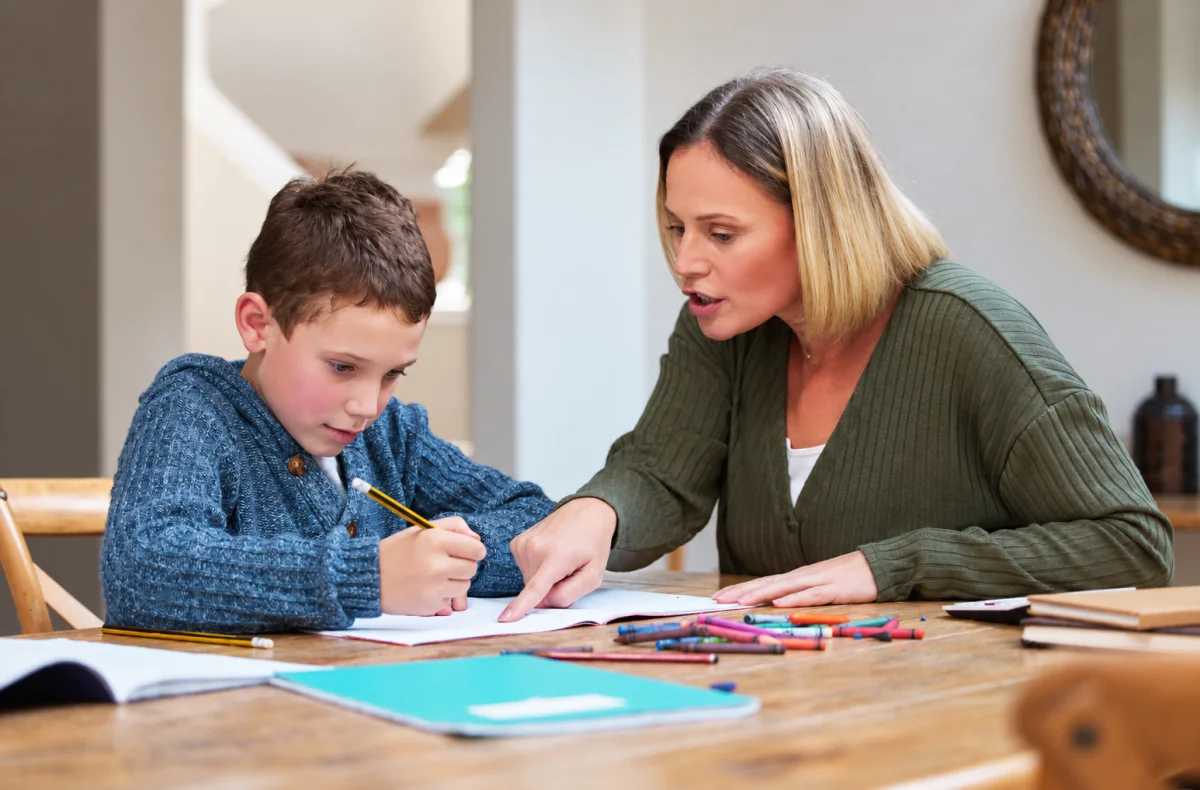 Una madre señala una parte del cuaderno mientras su hijo escribe con lápiz en la mesa de casa.