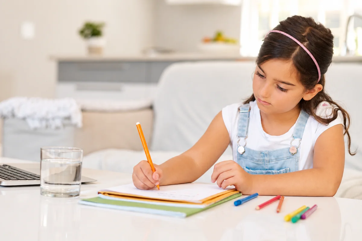 Una niña escribe en su cuaderno junto a un vaso de agua, una computadora y lápices de colores.