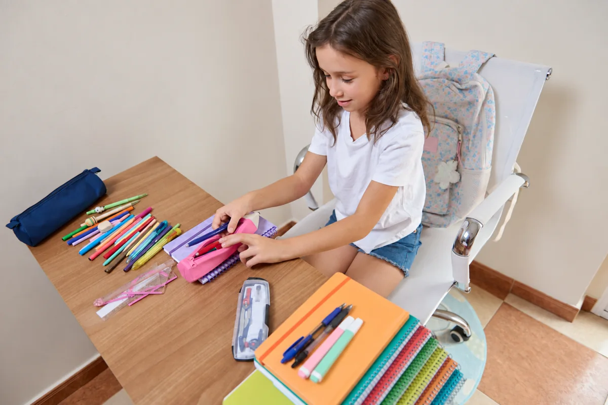 Una niña organiza lápices en un estuche rosa, con cuadernos y mochila preparados junto a su escritorio.