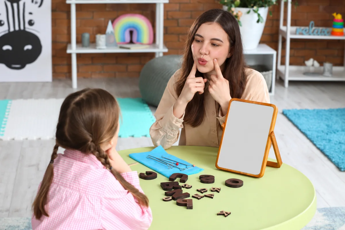Profesional y niña realizan un ejercicio de articulación frente a un espejo, con letras y materiales sobre la mesa.