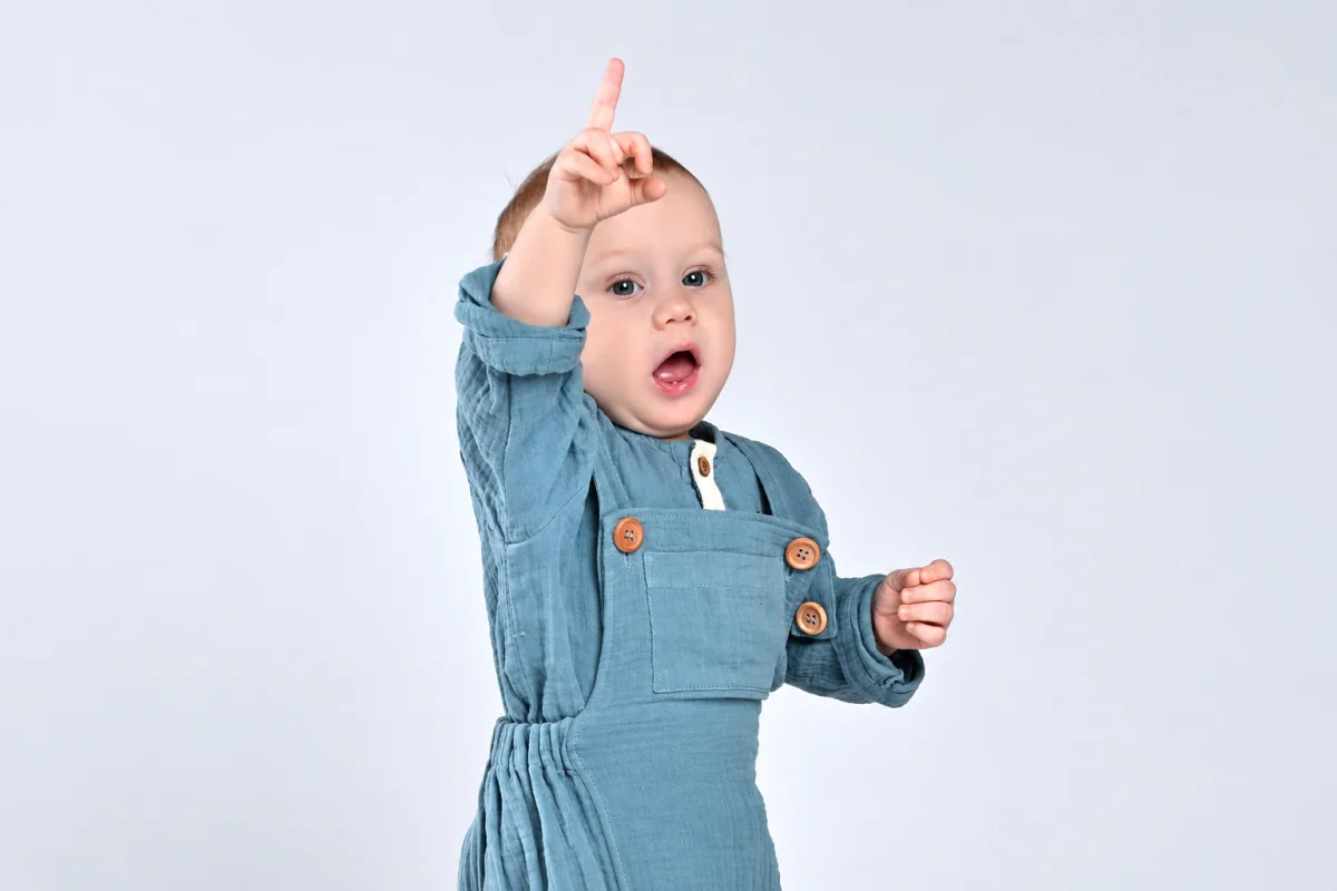 Niño pequeño con overol azul señala hacia arriba con el índice y mantiene la boca abierta.