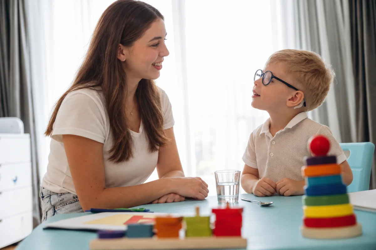 Una profesional conversa con un niño junto a un vaso y una cuchara sobre la mesa.