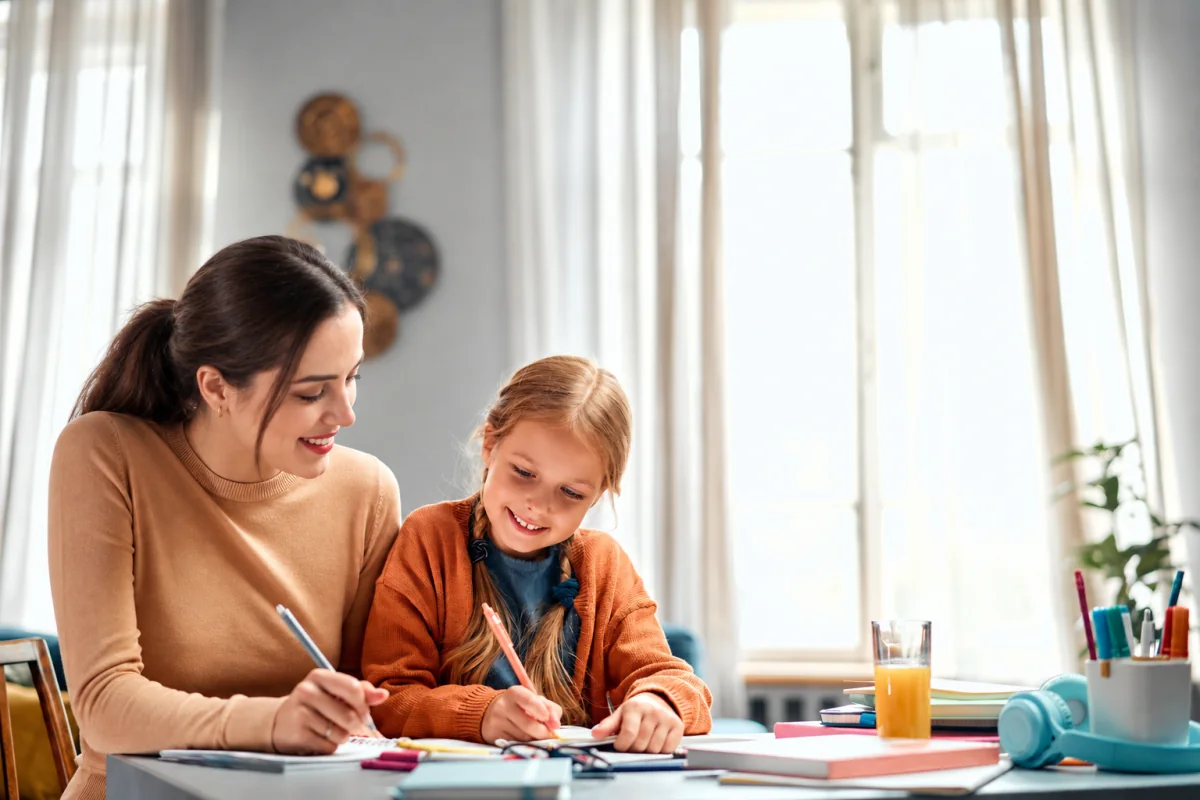 Adulta y niña escriben juntas en cuadernos sobre una mesa con libros, lápices y materiales escolares.